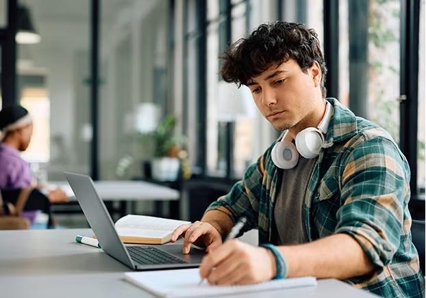 Student with headset using a laptop, pen, and paper to complete online coursework.